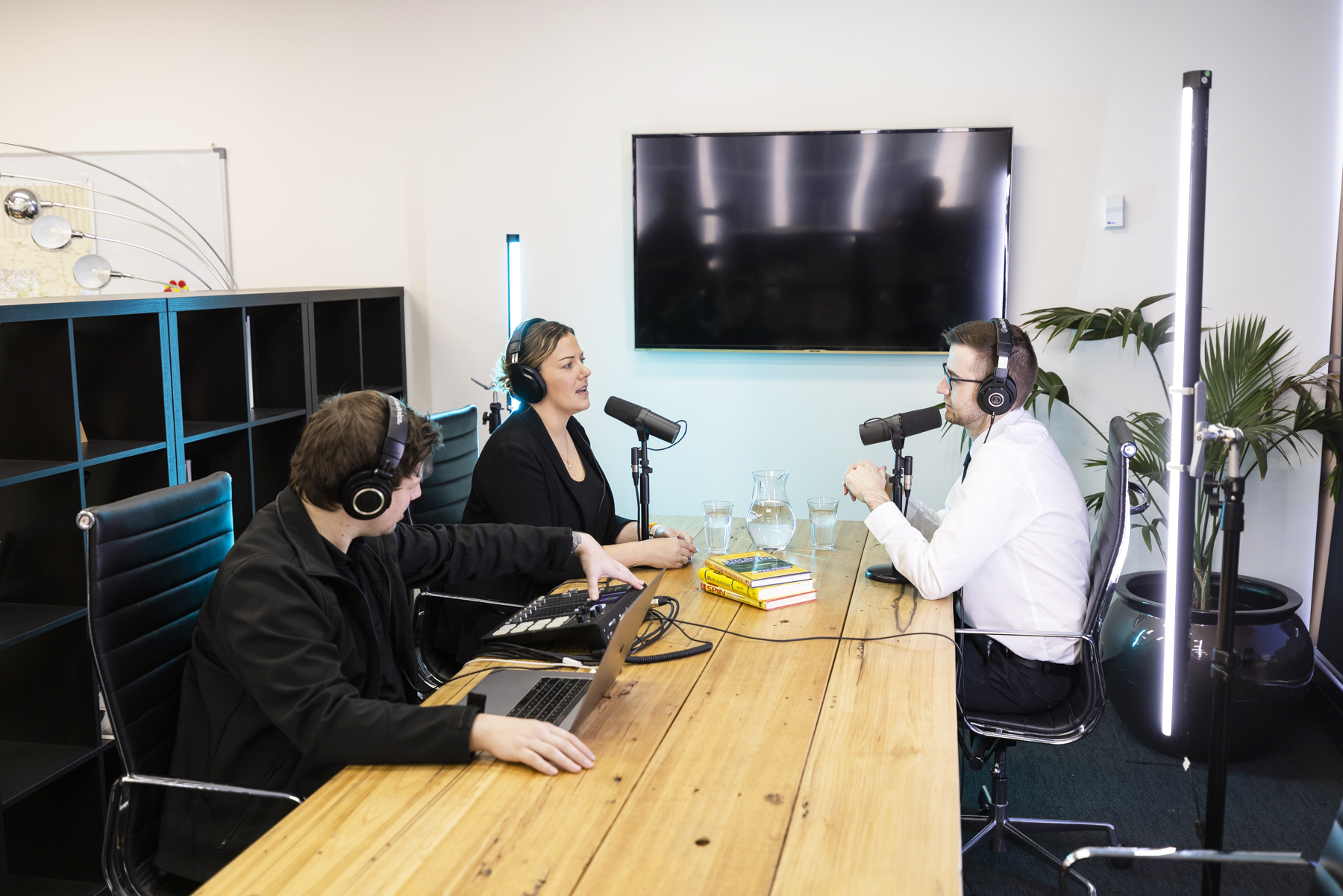 3 people sitting around a table recording a podcast, 2 are talking into microphones with headsets on