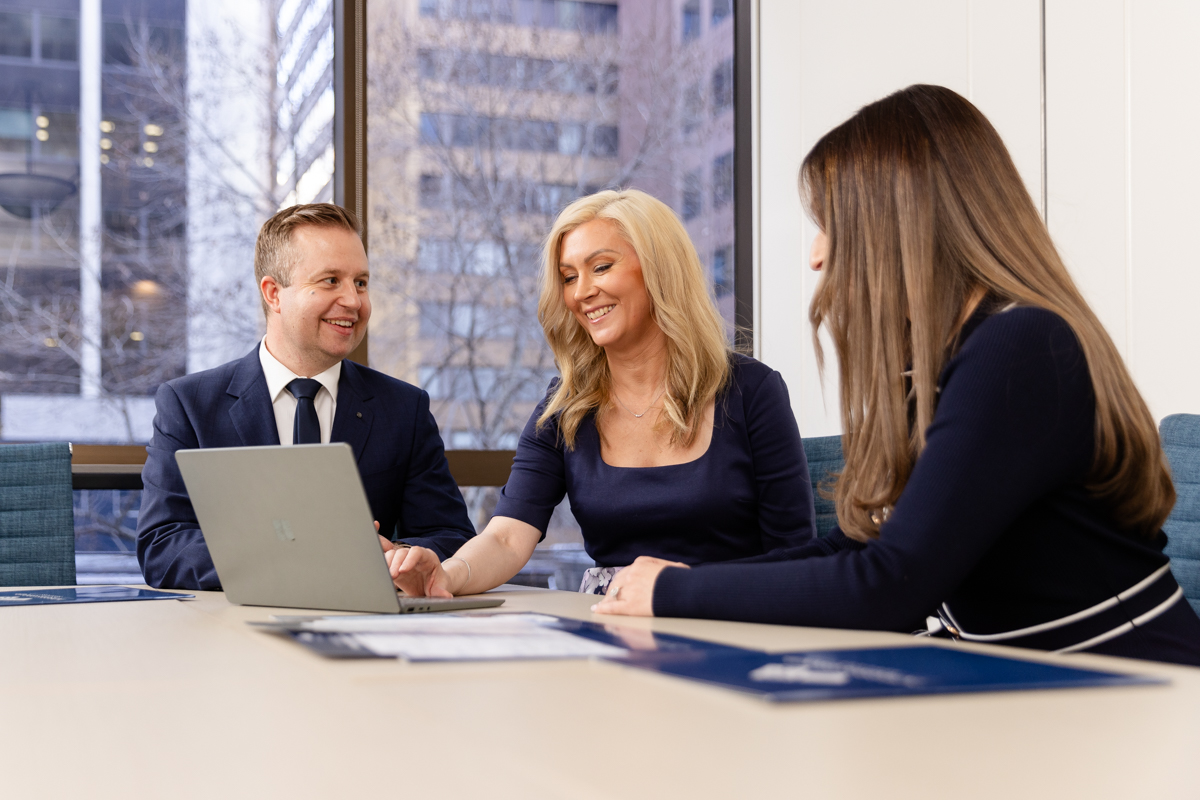 Three people sitting at boardroom table, looking at the laptop of the woman in the middle who is smiling